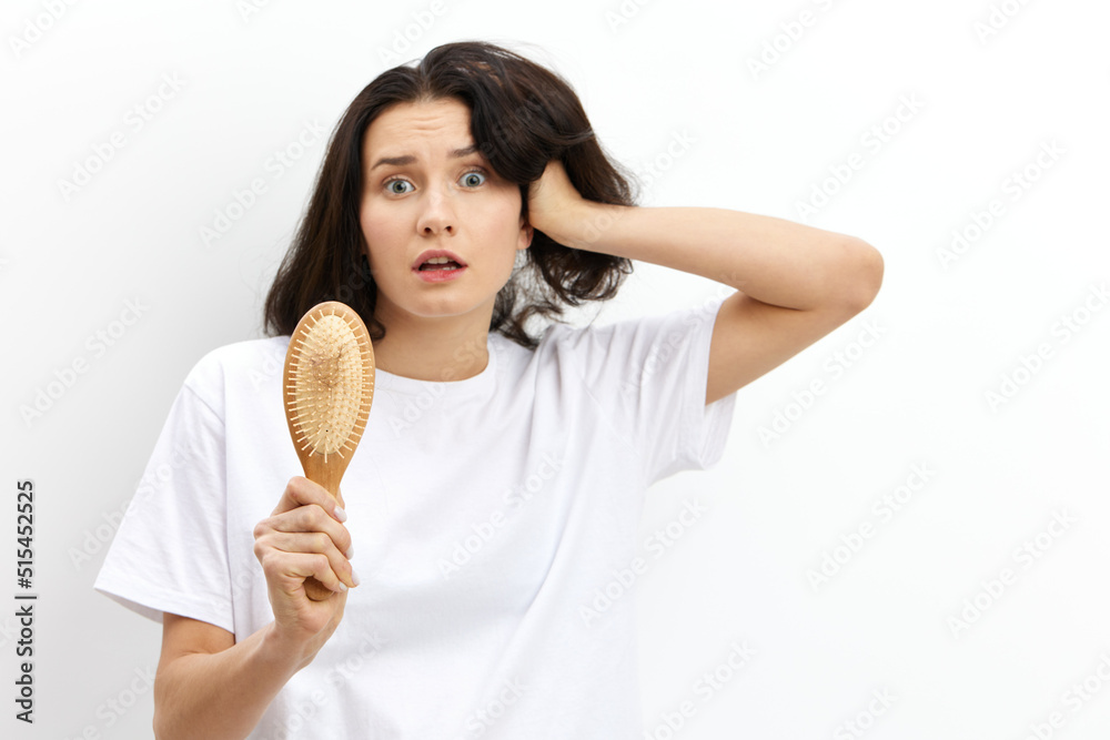 a sad, upset, puzzled woman with long black hair stands in a white T-shirt on a light background and holding her hand on her head in panic looks at her wooden comb a sad, upset, puzzled woman with long black hair stands in a white T-shirt on a light background and holding her hand on her head in panic looks at her wooden comb