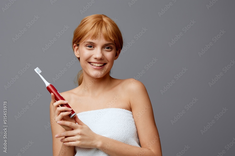 Horizontal studio shot. An emotional woman in a towel with light clean skin, with red silky hair gathered in a ponytail on a gray background with a toothbrush in her hands shows a like. Horizontal studio shot. An emotional woman in a towel with light clean skin, with red silky hair gathered in a ponytail on a gray background with a toothbrush in her hands shows a like.