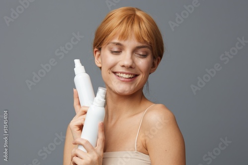 Close-up photo.Horizontal studio shot. An emotional woman in a white T-shirt with light, clear skin, with shiny red hair gathered in a ponytail stands on a gray background with two white bottles in photo