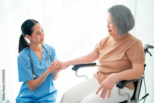 Asian nurse or doctor sit on floor and hold hands of senior woman sit in front of curtain in living room.