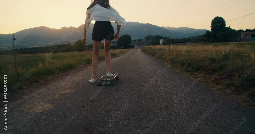 Young carefree and stylish adult woman ride on skate longboard at sunset along meadow. Trendy teenager girl skateboarding with mountain view. Concept of youth and freedom on countryside