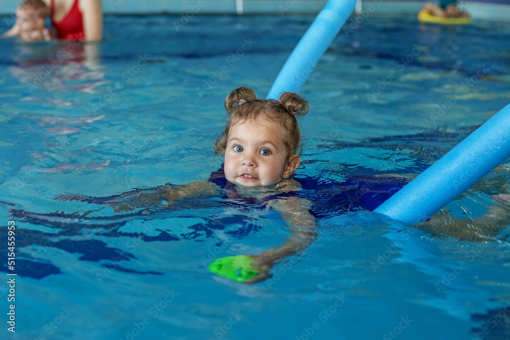 Little curly toddler girl learns to swim in swimming pool using foam stick.