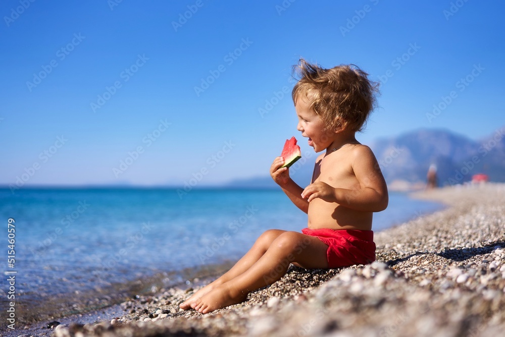 Adorable Young Toddler Eats Delicious Ripe Watermelon by the Ocean on Beautiful Beach During the Summer Adorable Young Toddler Eats Delicious Ripe Watermelon by the Ocean on Beautiful Beach During the Summer