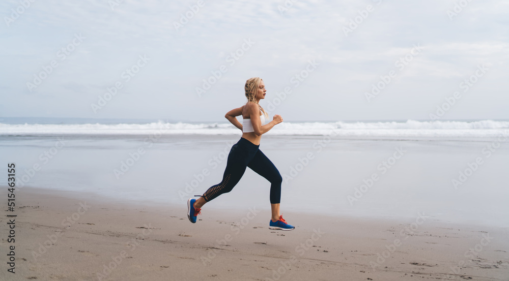 Full length view, Caucasian female with athletic sporty body running on seashore while training outdoors. Young woman working out and enjoying healthy lifestyle at nature. Runner moves on ocean beach