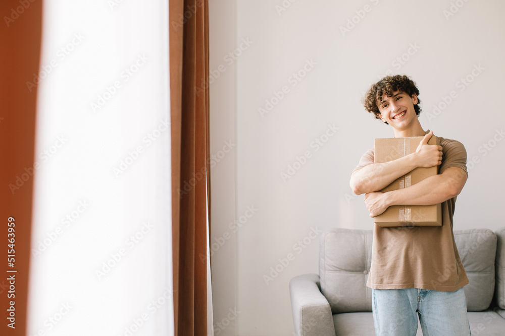 Shopping And Delivery. Curly Young Man Customer Holding Cardboard Box Posing Smiling To Camera Standing At Home. Excited Male Buyer Hugging Delivered Parcel. Shopaholism Concept Shopping And Delivery. Curly Young Man Customer Holding Cardboard Box Posing Smiling To Camera Standing At Home. Excited Male Buyer Hugging Delivered Parcel. Shopaholism Concept