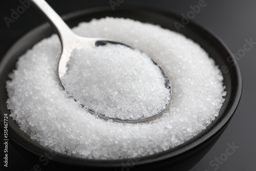 Granulated sugar and spoon in bowl on black table, closeup