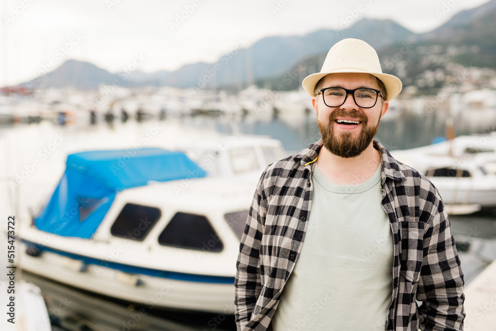 Handsome man wearing hat and glasses near marina with yachts. Portrait of laughing man with sea port background with copy space Handsome man wearing hat and glasses near marina with yachts. Portrait of laughing man with sea port background with copy space