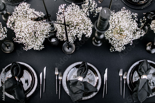 Murais de parede Top view of black style banquet table with a black tablecloth, silver plates, napkins, cutlery, black candles, and white flowers