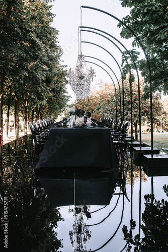 An outdoor banquet table decorated in black with crystal chandeliers on top of a mirrored floor. Reflection on the floor, black and white decor. Front view.