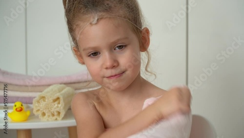 Portrait of little girl with hair net wipes wet face with soft fresh terry towel in bathroom. Happy child after bathing. Baby care.