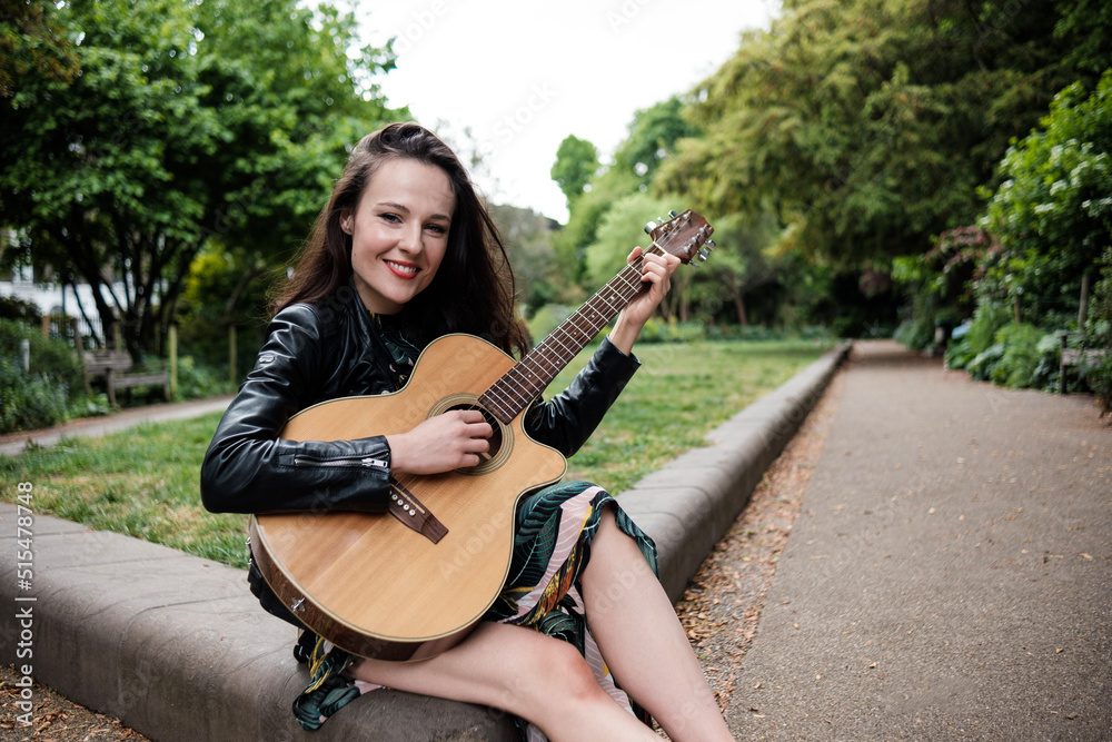 Obraz premium Portrait of smiling young woman playing guitar in a park and looking at camera.