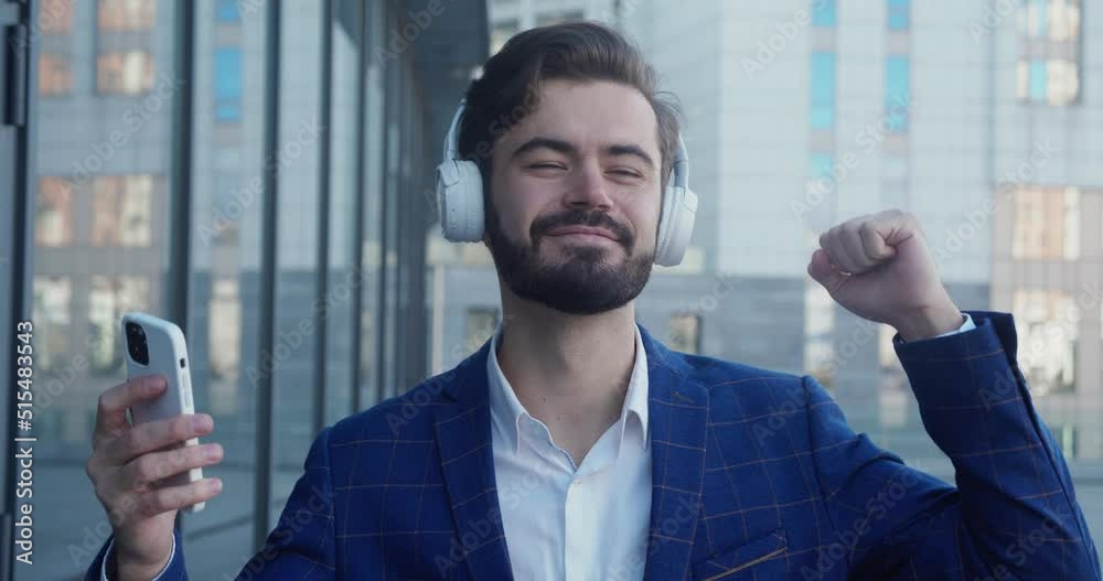 Caucasian happy beautiful man in suit in headphones dansing on street and listening to music outdoors. Joyful male in good mood enjoying songs in town. Close up portrait. Leisure concept.