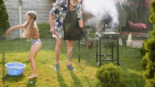 Old man is grilling in the backyard while children are splashing with water balloons on a hot summer day.