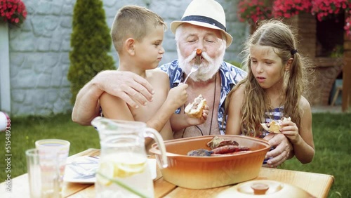 Slow motion shot of little boy and girl sitting on grandfather's lap and feeding him fresh meat from the grill in the yard