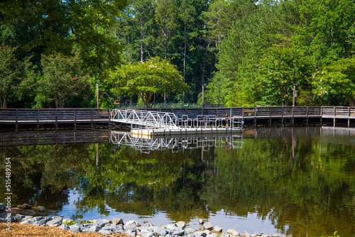 a gorgeous summer landscape at the lake with a brown wooden bridge and dock with a metal hand rail surrounded by lush green trees reflecting off the water at Dupree park in Woodstock Georgia USA photo