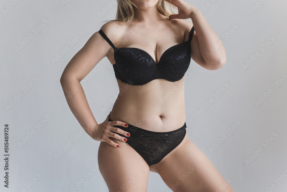 A young woman with curves and cellulite poses in black lingerie on a gray  background without