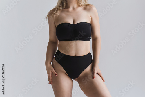 A young woman with curves and cellulite poses in black sports underwear on a gray background without a face. Plus size model posing in lingerie.