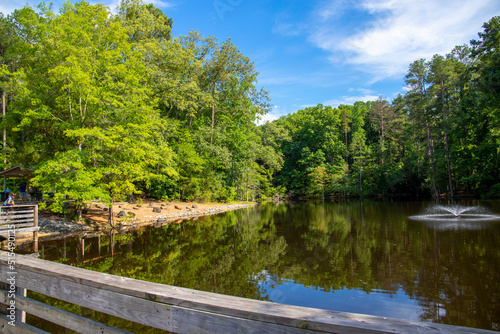 a gorgeous summer landscape in the park with a lake and a water fountain and a brown wooden bridge surrounded by lush green trees, grass and plants with blue sky and clouds at Dupree park in Woodstock photo