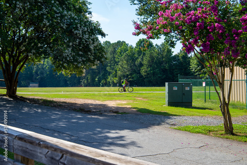 a man wearing a yellow shirt and a black helmet and backpack riding a bike in the park with a gorgeous summer landscape surrounded by lush green trees, grass and plants at Dupree Park in Woodstock photo