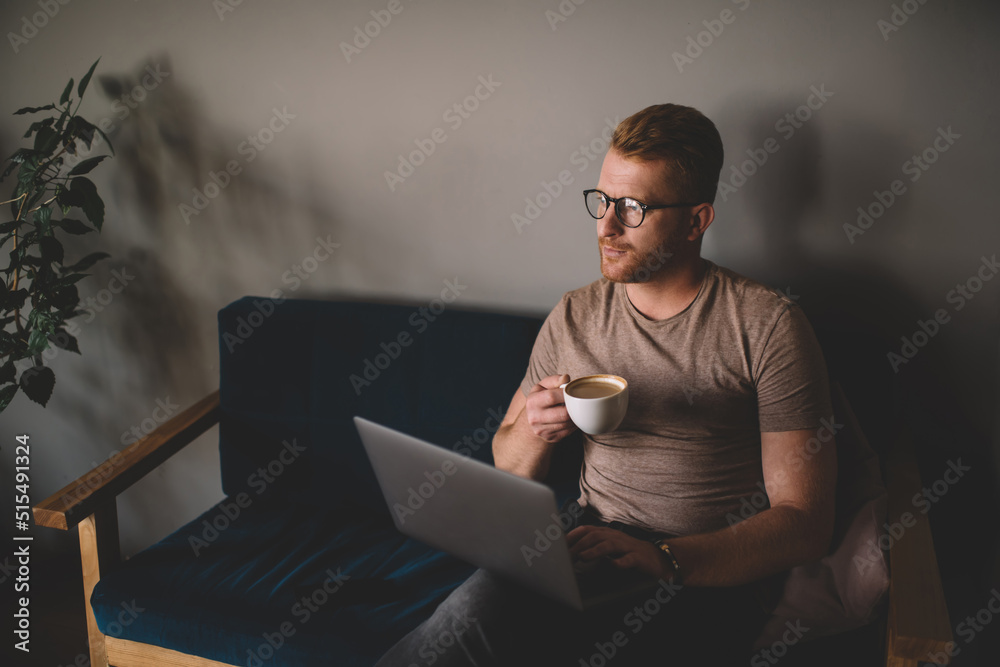 Redhead man working on freelance project with laptop computer in coworking office indoors. Digital nomad reading news on website while sitting with netbook and drink coffee. Student learning online
