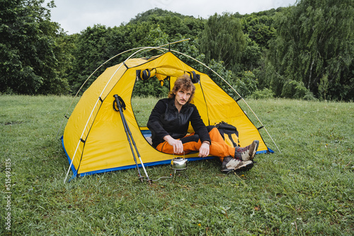 Summer vacation in the mountains in nature guy sitting in a tent in a campsite, hiking through the forests alone, yellow tent, survival in the taiga, trekking on foot in the mountains