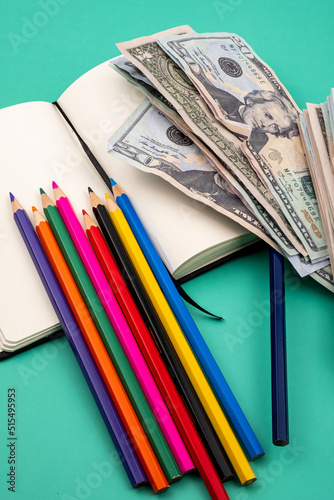 Several colored pencils on top of a notebook and a pile of dollars, on a green table with a white background. Back to school is very expensive.