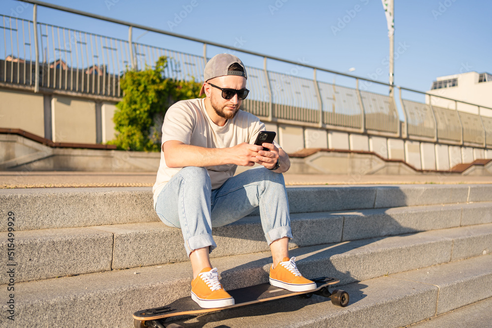 man using mobile phone while sitting with skateboard at city street.Young skater guy typing message by cellphone outside. 