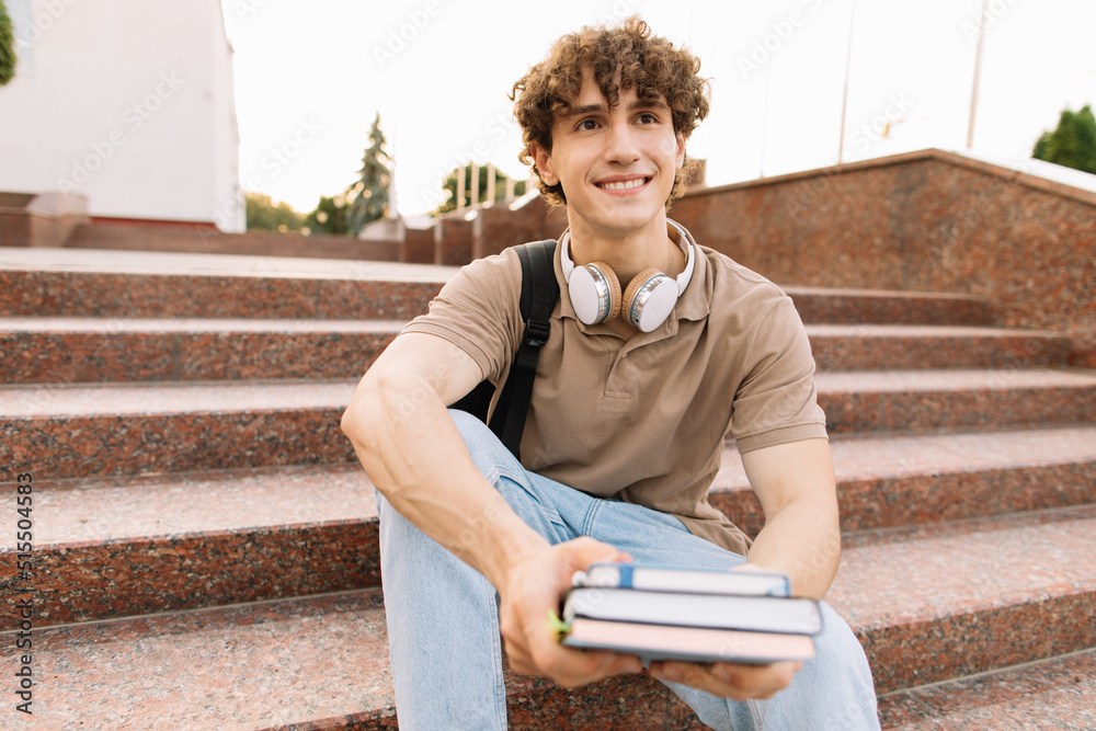 Obraz premium Attractive male student sits on the steps of the university and reads something, preparing for exams