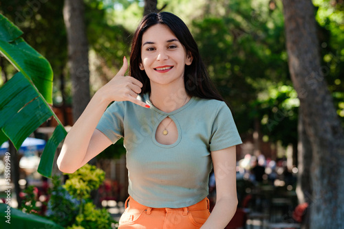 Young brunette girl smiling happy wearing turquoise t-shirt standing on city park, outdoors smiling doing phone gesture with hand and fingers like talking on the telephone. Communicating concepts.