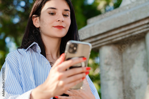 Young brunette woman wearing blue striped shirt on city park, outdoors looking at the phone screen and using phone. Messaging with friends, watching video or scrolling on social media.