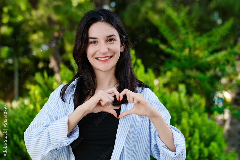 Cheerful brunette woman wearing blue striped shirt on city park, outdoors showing heart gesture with two hands and looking at camera. Heart shape with hands. Cheerful brunette woman wearing blue striped shirt on city park, outdoors showing heart gesture with two hands and looking at camera. Heart shape with hands.
