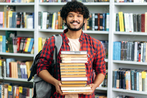 Happy smart indian or arabian guy, mixed race male, university student, stands in the library against the background of bookshelves, holds a lot of books in his hands, looks at camera, smiles friendly