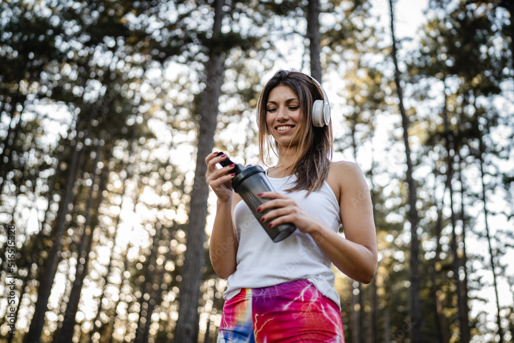 One woman happy caucasian female standing in the forest or park in woods with supplement shaker and headphones during training in nature taking a brake water drink and hydration real people copy space