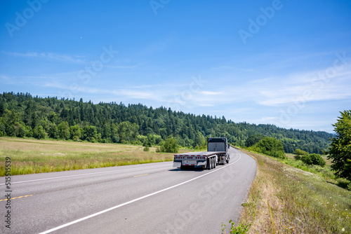 Classic black big rig semi truck with empty flat bed semi trailer driving on the winding summer road to warehouse for the next load