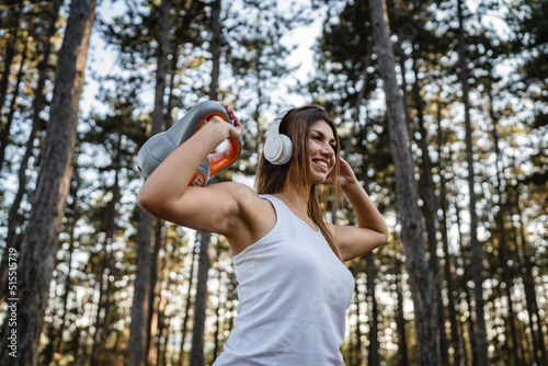 One woman adult caucasian female athlete using kettlebell girya weight during training in the forest park woods in summer day with headphones happy smile brunette health and fitness concept copy space © Miljan Živković