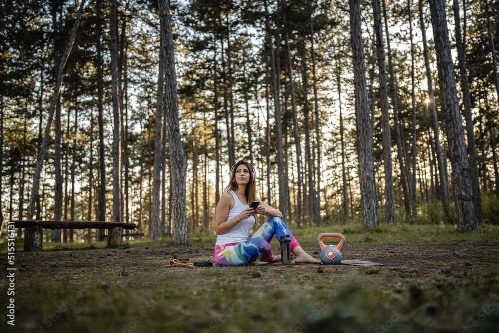 One woman adult caucasian female using mobile phone while taking a brake during training in the forest woods or park in summer day with kettlebell equipment listen music on earphones and mobile app One woman adult caucasian female using mobile phone while taking a brake during training in the forest woods or park in summer day with kettlebell equipment listen music on earphones and mobile app