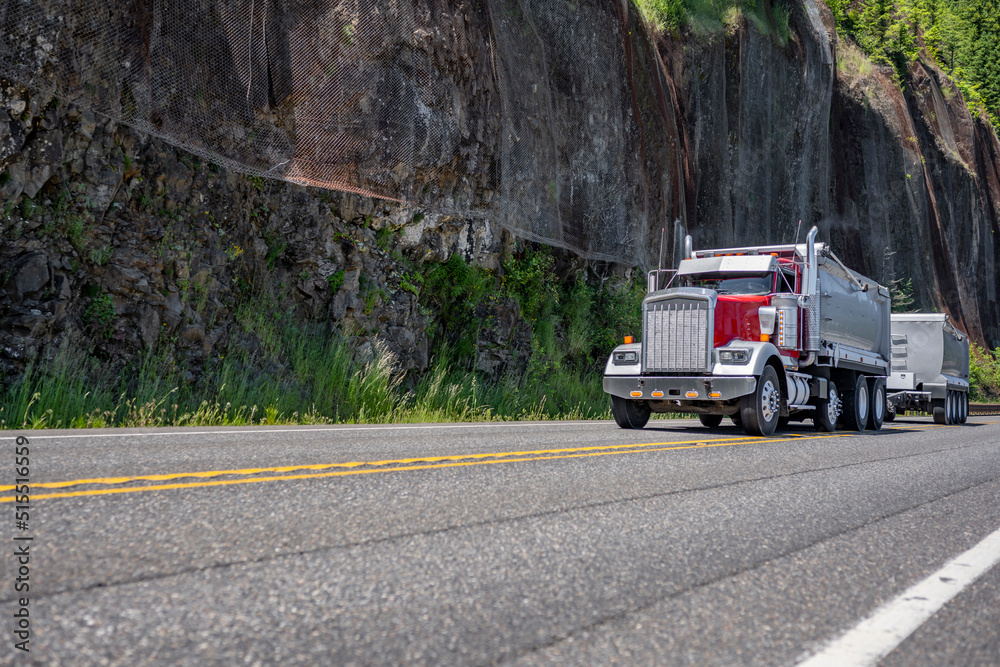 Red tipper rig semi truck with two tip trailers driving on the mountain road with rock cliff wall on the side