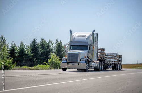 White classic big rig semi truck tractor transporting fastened lumber cargo on step down semi trailer running on the flat road with trees on the side