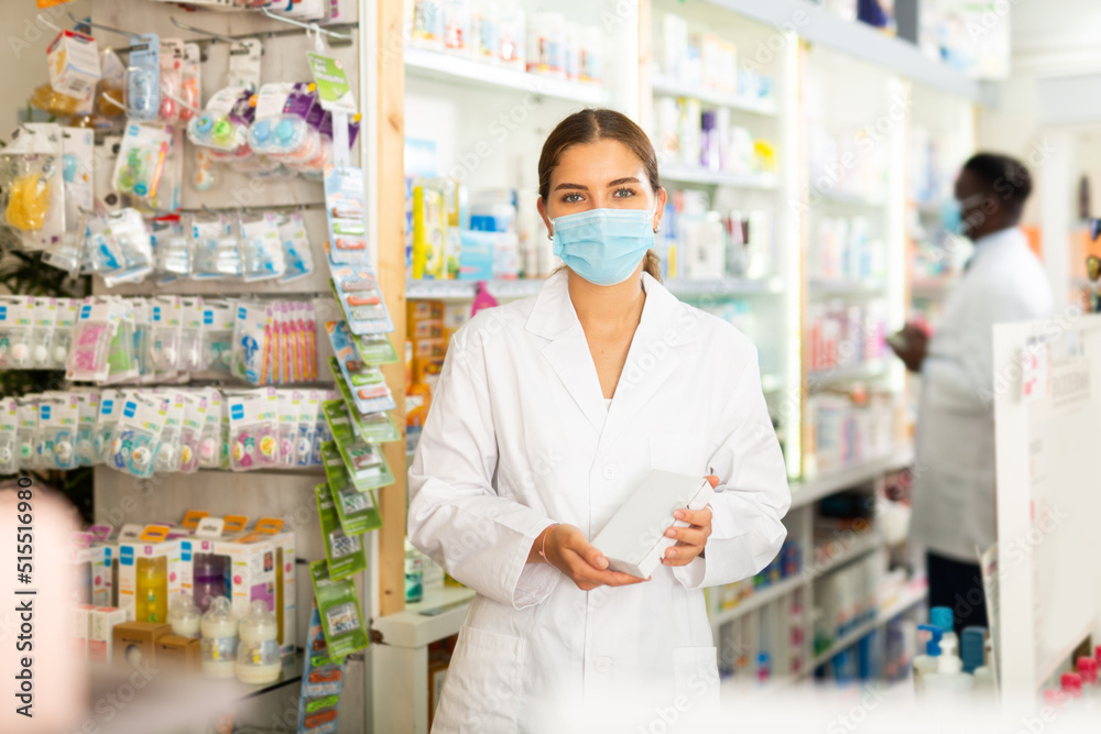 Portrait of a young female pharmacist in a protective mask, working in a pharmacy during the pandemic, standing in the ..trading floor and demonstrates in her hands a goods that was recently put up