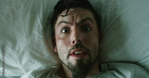 A man suffering from insomnia has a nightmare in bed at night. A scared man gets frighted and looks terrified after having a bad, horrific dream. Guy wakes up and panics while lying in a dark room