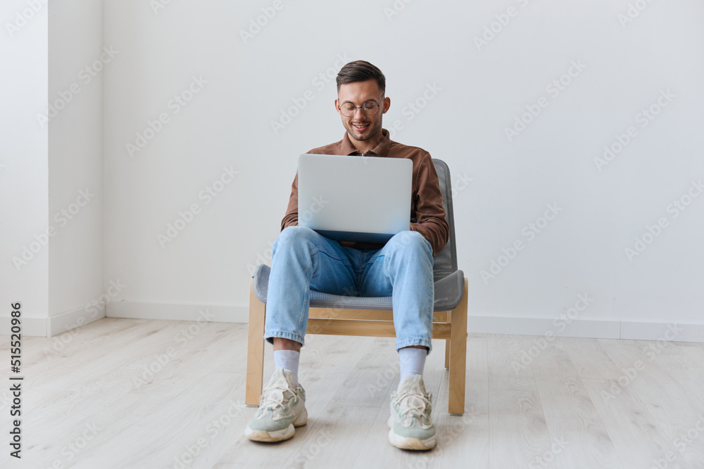 Smiling happy young tanned man in eyewear hold laptop on knees chatting with friends typing message looks at screen sitting on chair at white home wall. Lottery winner offer. Copy space