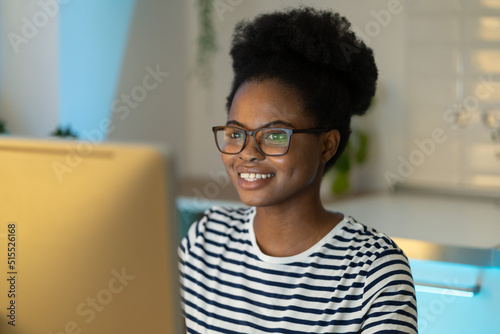 Cheerful joyful black girl remote worker wearing eyeglasses looking at computer monitor while working at home office  happy African woman social media specialist working online. Future of remote work