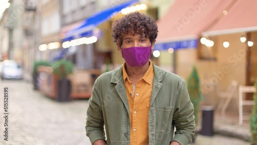 Camera approaching mixed-race happy guy in fask standing in town smiling at camera Multiracial young joyful stylish man on street with cafeterias and restaurants during covid pandemic, tourism concept photo