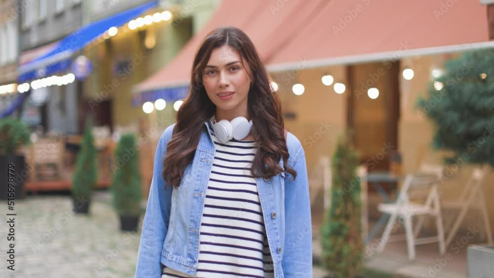 Portrait of beautiful Caucasian woman with headphones standing in city center in good mood smiling at camera. Positive young female tourist in downtown with happy face expression. Leisure, tourism Portrait of beautiful Caucasian woman with headphones standing in city center in good mood smiling at camera. Positive young female tourist in downtown with happy face expression. Leisure, tourism