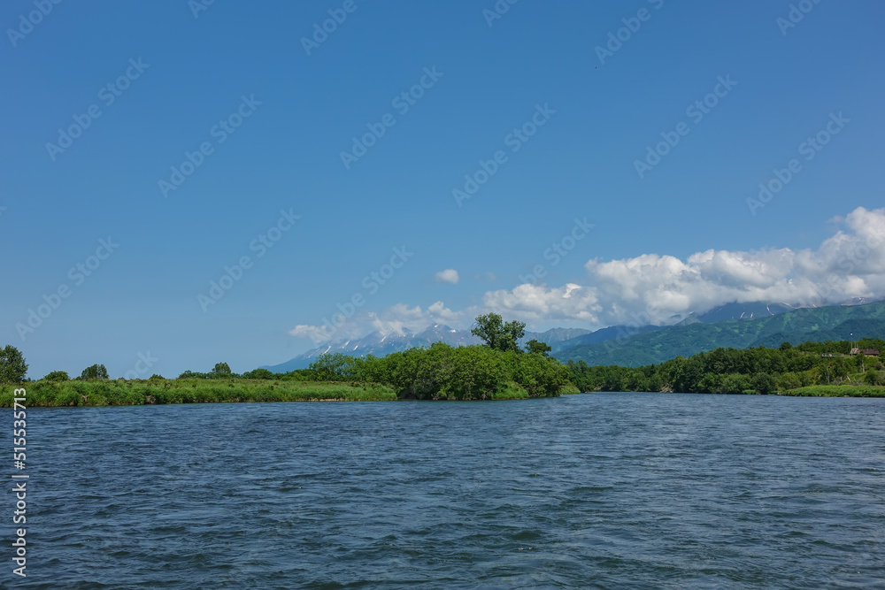 The bed of the blue river bends between the green banks. A picturesque mountain range against the background of azure sky and clouds. Copy space. Kamchatka. River Bystraya The bed of the blue river bends between the green banks. A picturesque mountain range against the background of azure sky and clouds. Copy space. Kamchatka. River Bystraya