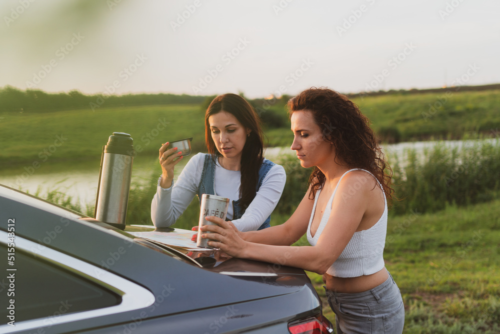 two girls are traveling on the roads by car, stopped at the side of the road and look at the map, drinking coffee from a thermos. Vacation concept two girls are traveling on the roads by car, stopped at the side of the road and look at the map, drinking coffee from a thermos. Vacation concept