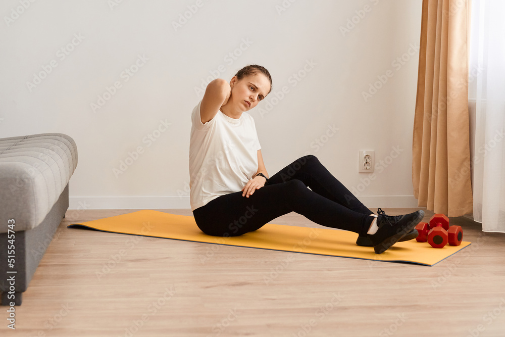 Fototapeta premium Portrait of beautiful woman sitting on yoga mat feeling neck pain while doing yoga and exercise at home, correct exercise technique and be careful, female wearing white t shirt and black leggins.