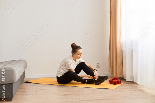 Full length portrait of unhealthy woman sitting on yoga mat doing exercise at home and injured her ankle, frowning face, feeling strong pain, posing in living room.