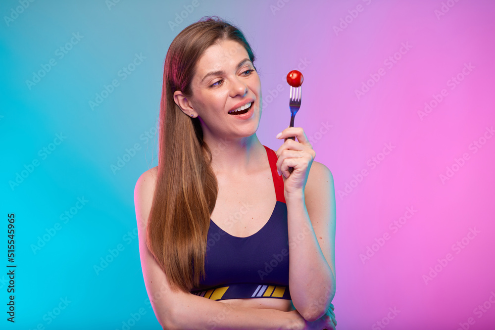 Smiling sporty woman in fitness bra holding red tomato on fork looking away. Female fitness portrait isolated on neon color background. Smiling sporty woman in fitness bra holding red tomato on fork looking away. Female fitness portrait isolated on neon color background.