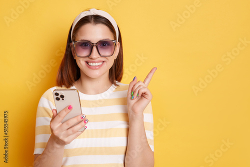 Portrait of happy smiling woman wearing sunglasses, striped T-shirt and hair band, using mobile phone and pointing finger at advertisement area, posing isolated over yellow background.